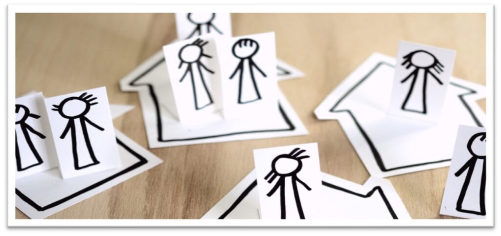 Das Bild zeigt acht  gemalte Figuren auf weissen Papierzetteln. Die Figuren unterscheiden sich durch unterschiedliche Frisuren. Zudem gibt es vier größere weisse Zettel, die die Umrisse von Häusern skizzieren. Die Figuren in unterschiedlichen Gruppenkonstellationen "in" den Häusern. Eine Figur steht ausserhalb.