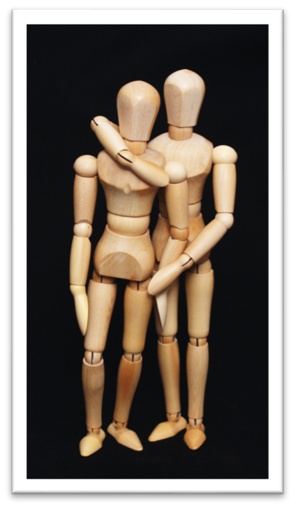 Zwei Holzfiguren stehen vor schwarzem Hintergrund. Die eine Holzfigur legt ihren Arm um die andere.
